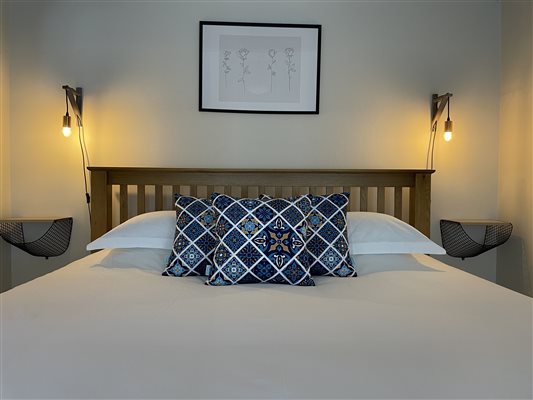 Super King size bed with white bed linen and three patterned cushions.  Above the bed is a picture.  On each side of the bed are wall lights, creating a warm soft light.
